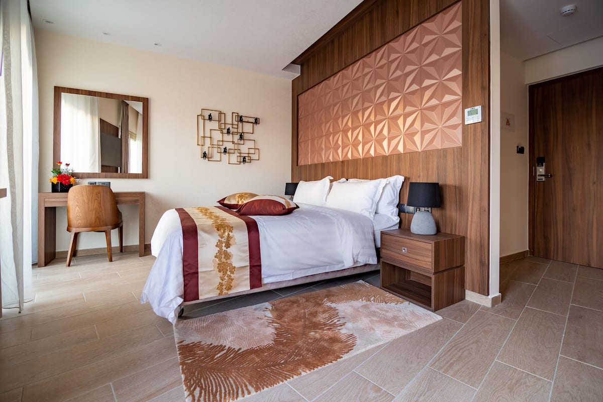 A modern hotel room with a large bed, a desk, a chair, a nightstand, and a wall-mounted mirror.