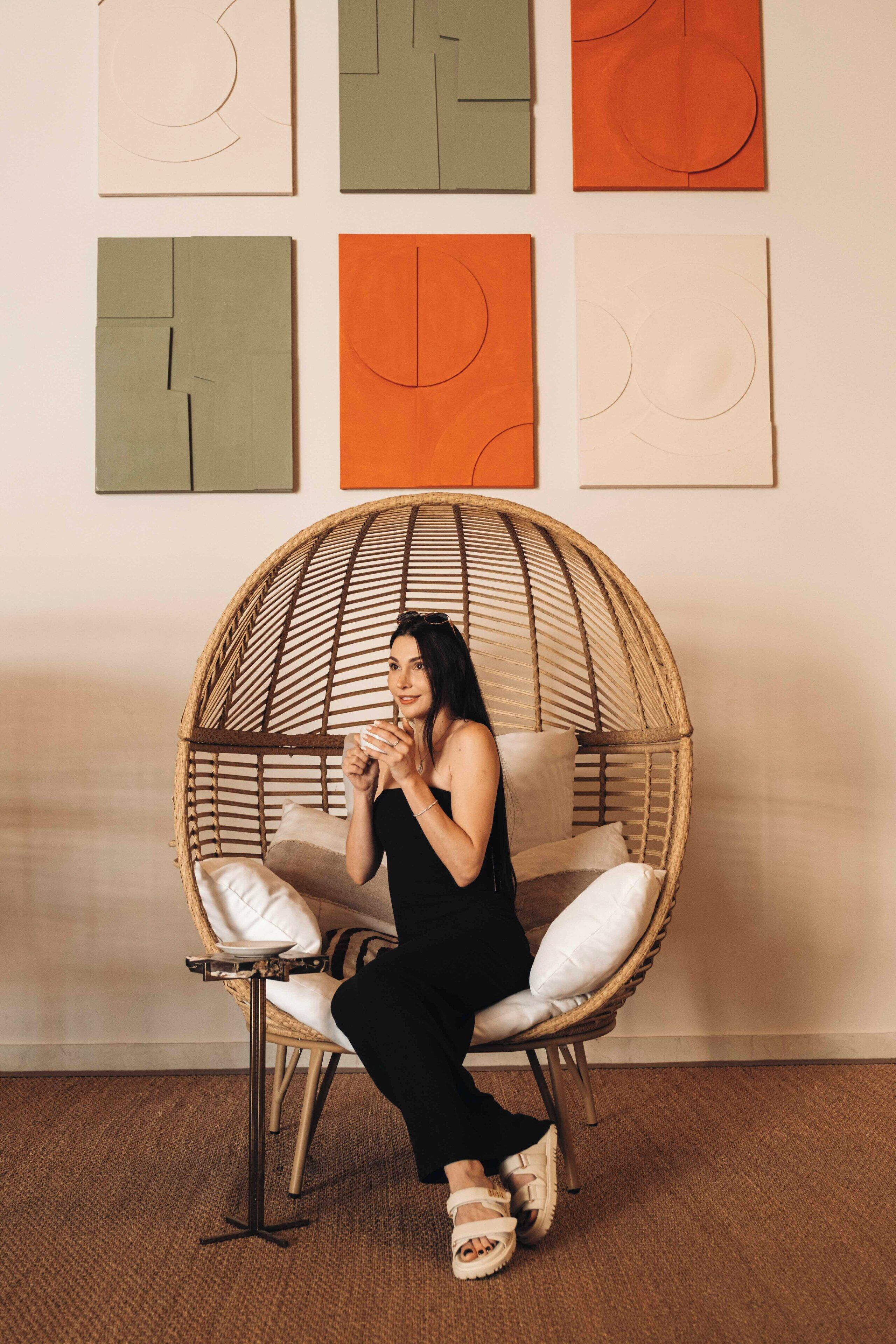 A person sitting in a wicker chair with a cushion, in a room with a plain wall and a carpeted floor.