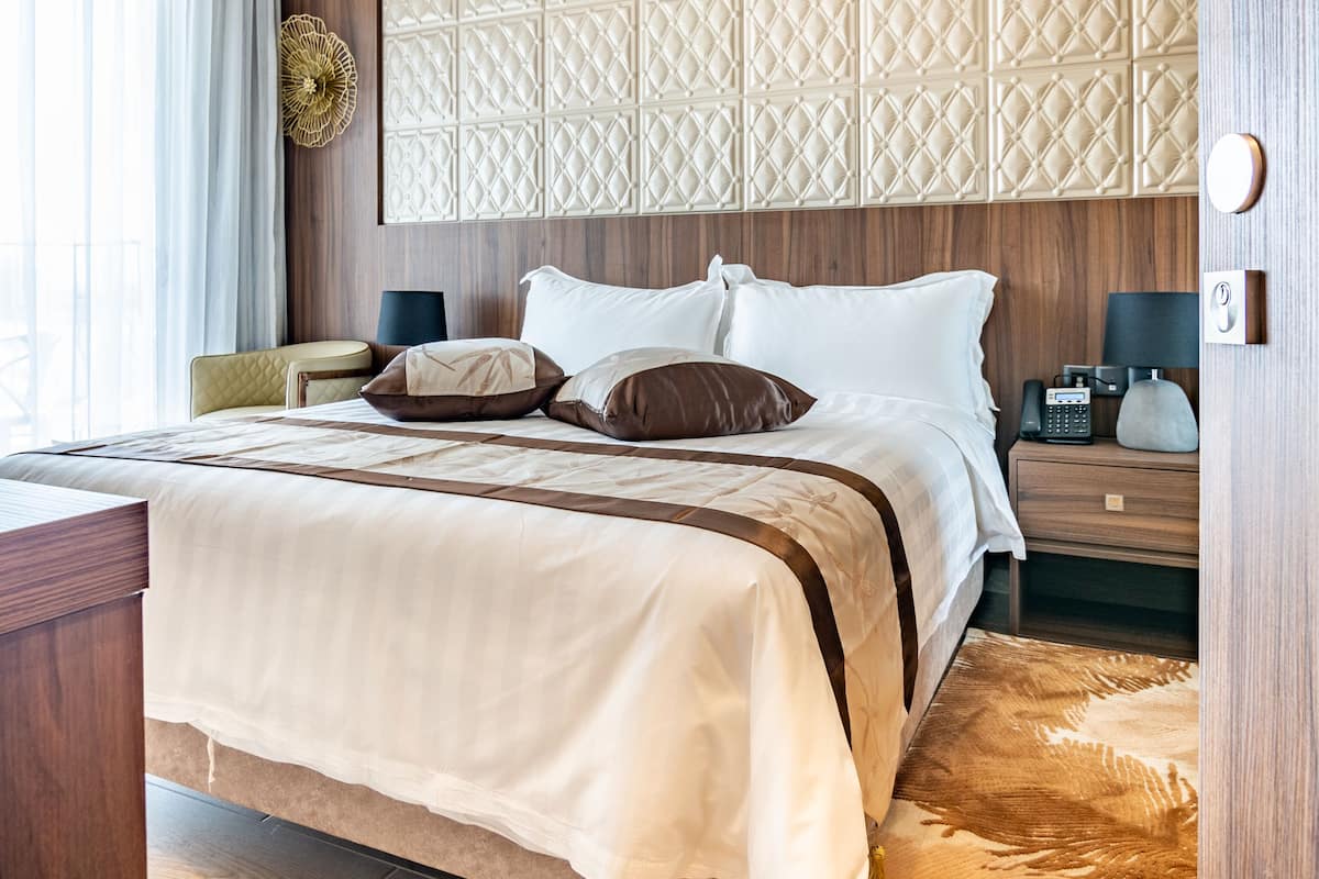 A hotel room with a wooden headboard, a large bed with white and brown bedding, a bedside table with a lamp, and a patterned rug.