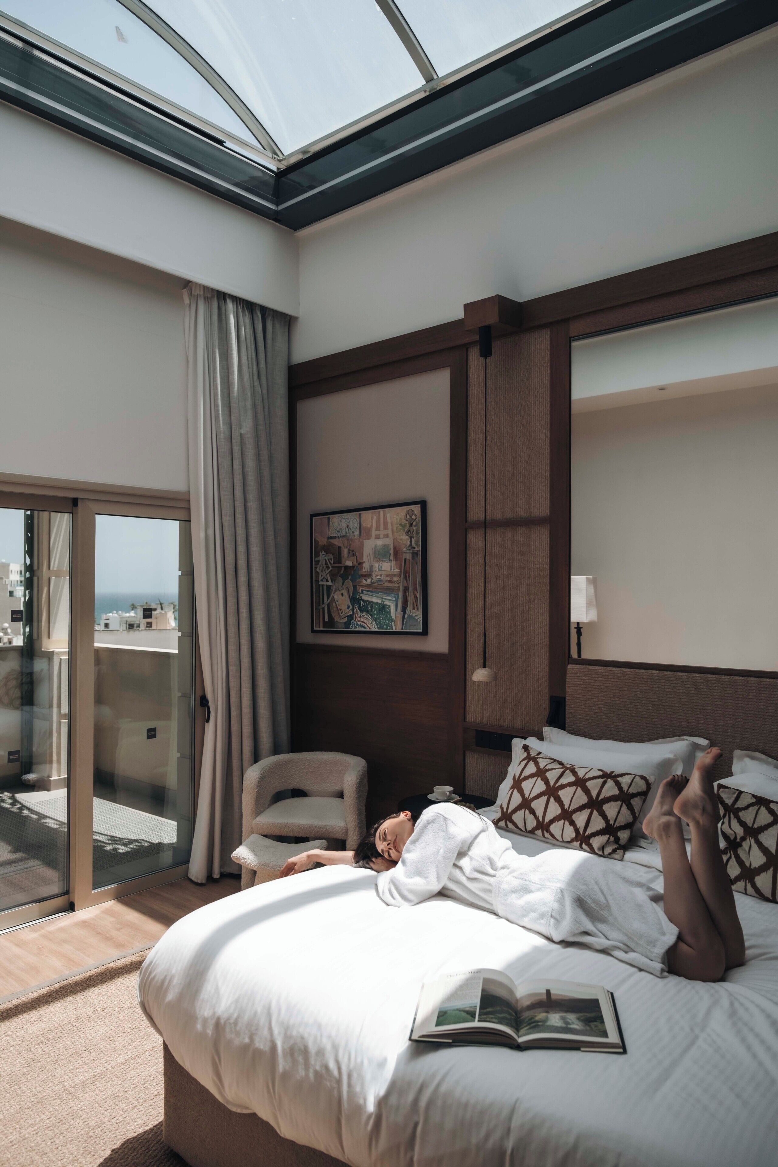 A hotel room with a large bed, a reading lamp, a painting on the wall, and a view of the city through a glass door.