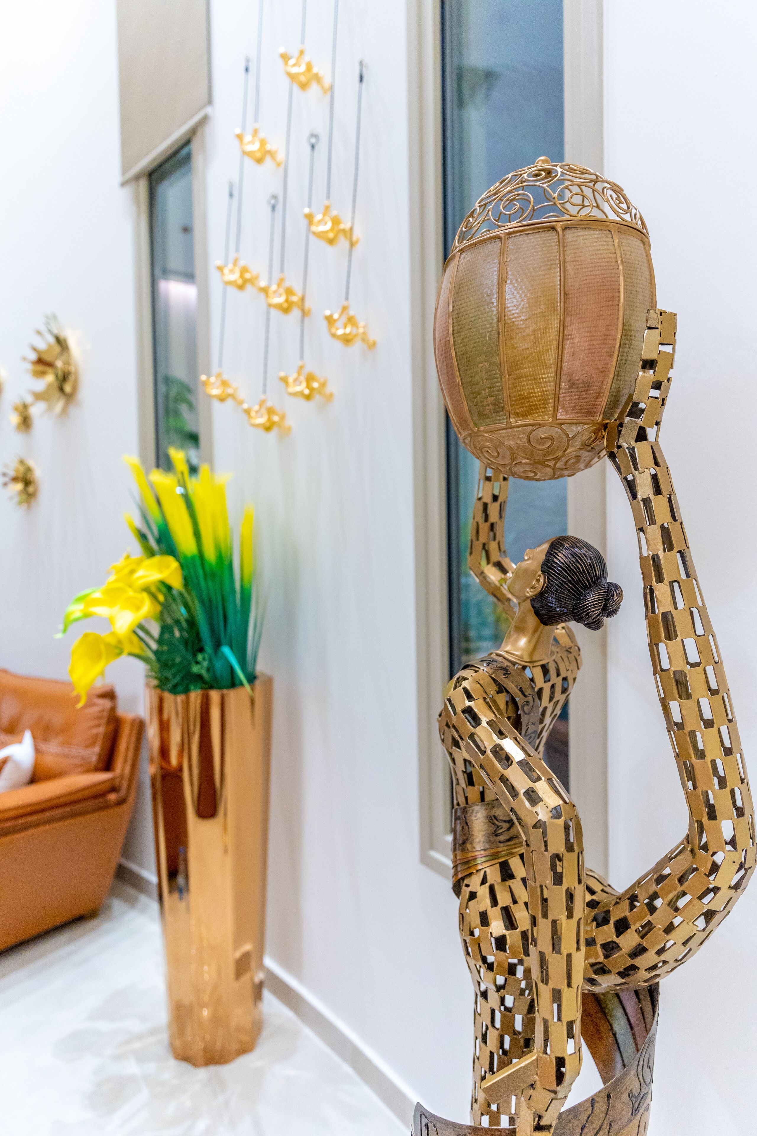 Escultura de uma mulher segurando um globo, um vaso dourado com flores amarelas e um sofá marrom em um quarto com paredes brancas e uma janela.