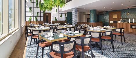 A spacious dining area with multiple tables and chairs, large windows, and hanging plants.
