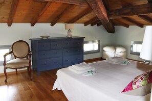 A bedroom with a wooden ceiling, a bed with white linens, a blue dresser, a chair, and a window with white curtains. - Stunning villa with private pool close to Rome in the Sabine countryside (Roccantica)
