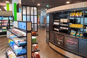 Un interior moderno con un mostrador que ofrece aperitivos y bebidas, estantes con productos envasados y una pantalla digital.