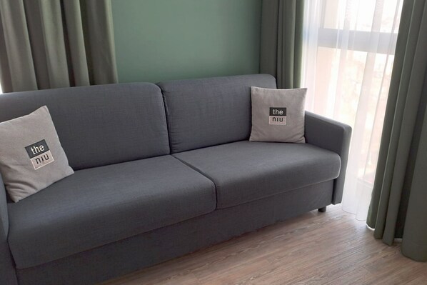 Un sofá gris con dos almohadones, uno con texto, en una habitación con suelo de madera y cortinas verdes.