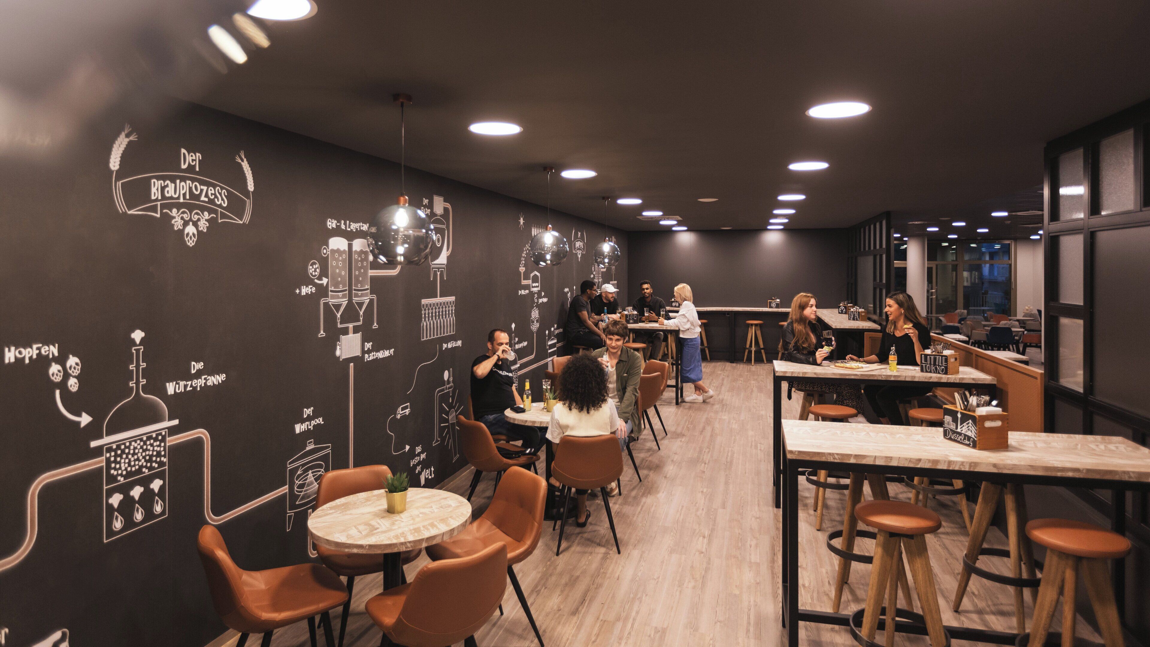 Een modern café met ronde tafels en oranje stoelen, met een muur waarop krijt tekeningen van brouwprocessen staan.