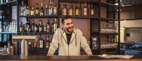 Uma área de bar com prateleiras de garrafas e copos, um balcão e uma pessoa atrás dele.