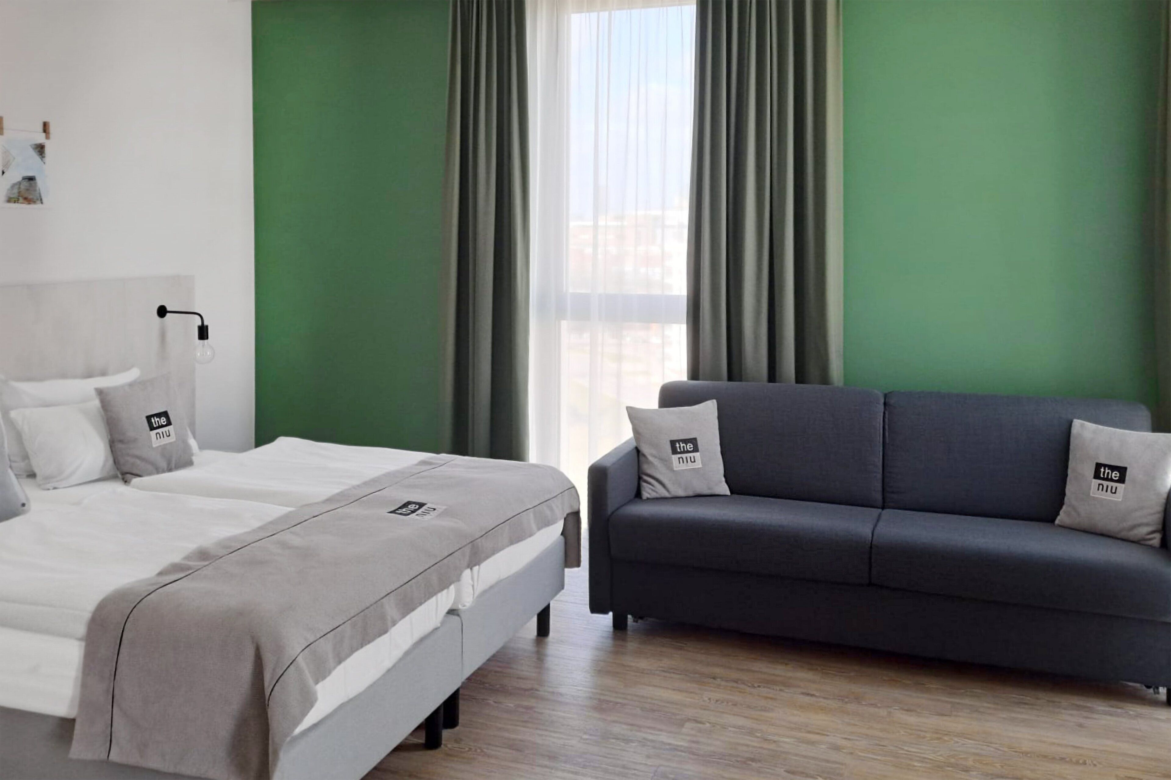 Habitación de hotel con cama, sofá, ventana con cortinas y lámpara fijada a la pared.