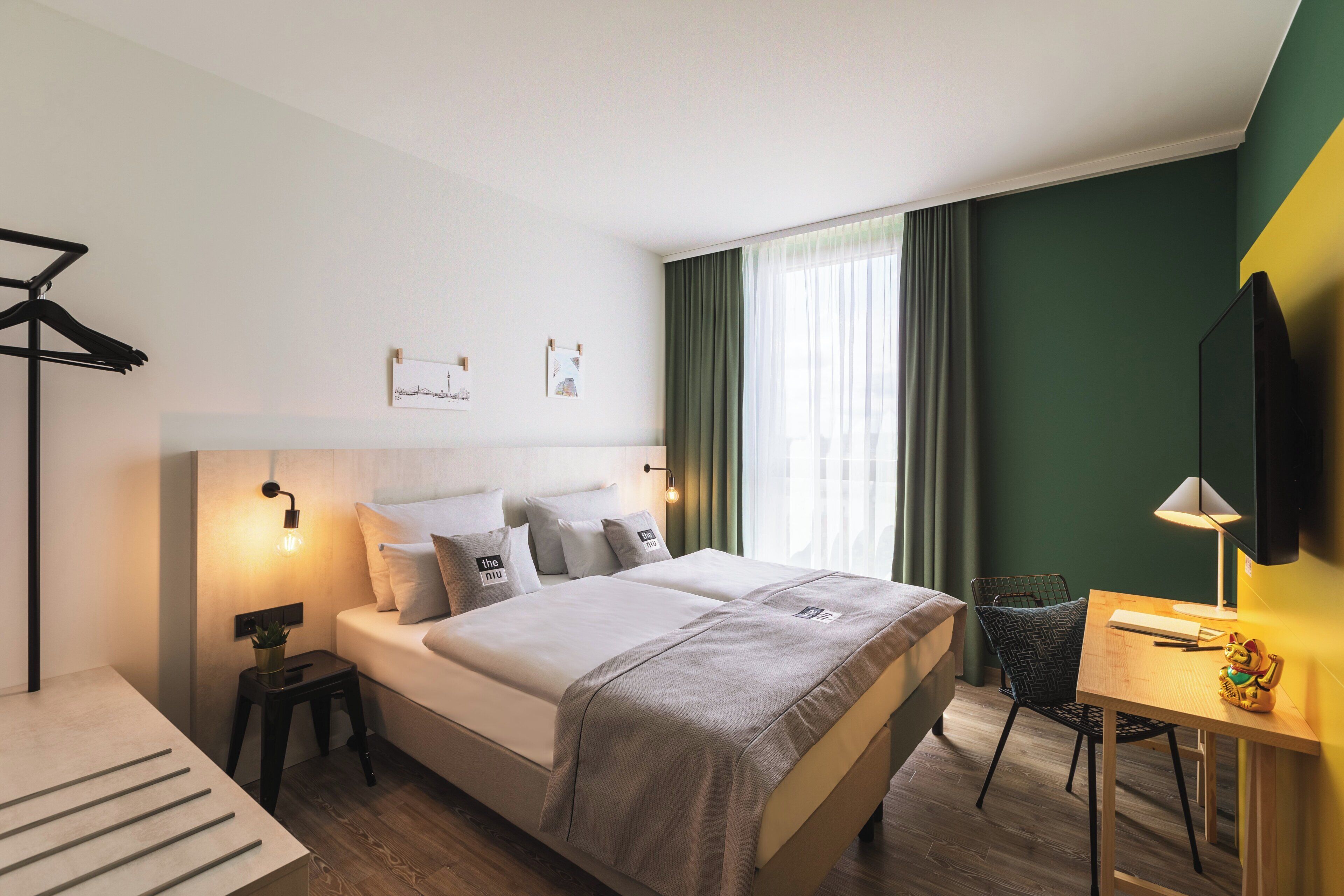 A modern hotel room with a large bed, bedside tables, a desk with a chair, a TV, and a window with curtains.