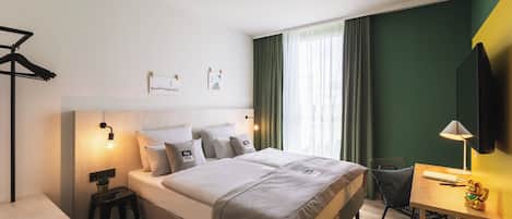 A modern hotel room with a large bed, bedside tables, a desk with a chair, a TV, and a window with curtains.