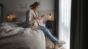 A person sitting on a bed in a hotel room, using a smartphone. The room has a modern design with a wooden headboard, two bedside lamps, and a view of the city through the window. - Holiday Inn - the niu, Tab Dusseldorf Main Station by IHG (Düsseldorf)