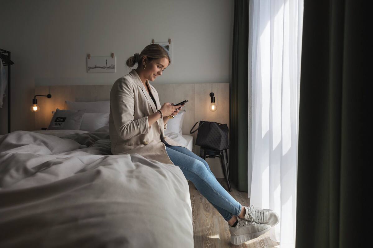 A person sitting on a bed in a hotel room, using a smartphone. The room has a modern design with a wooden headboard, two bedside lamps, and a view of the city through the window.