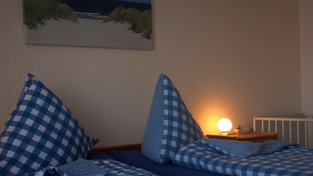 A bedroom with a checkered bedspread, pillows, a bedside table with a lamp, and a painting on the wall. - Vacation apartment in Haus Borkum 1 upper floor plus visitor's tax (Dornum)