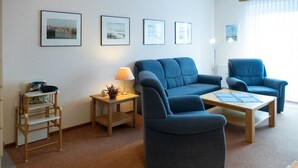 A living room with a blue sofa, two armchairs, a wooden coffee table, a side table with a lamp, and framed pictures on the wall. - Vacation apartment in Haus Borkum 1 upper floor plus visitor's tax (Dornum)