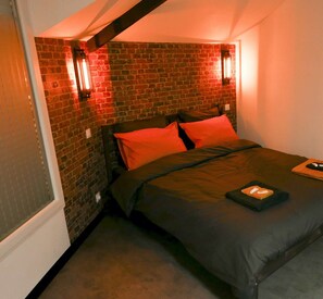 Un dormitorio con pared de ladrillo, una cama con ropa oscura y dos lámparas fijadas a la pared.