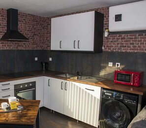 Una kitchenette compacta con pared de ladrillo, extractor de aire negro, mueble blanco, microondas rojo y lavadora.