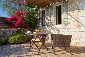 Relax in a serene outdoor setting with a wicker chair and small table, surrounded by lush greenery and vibrant flowers.