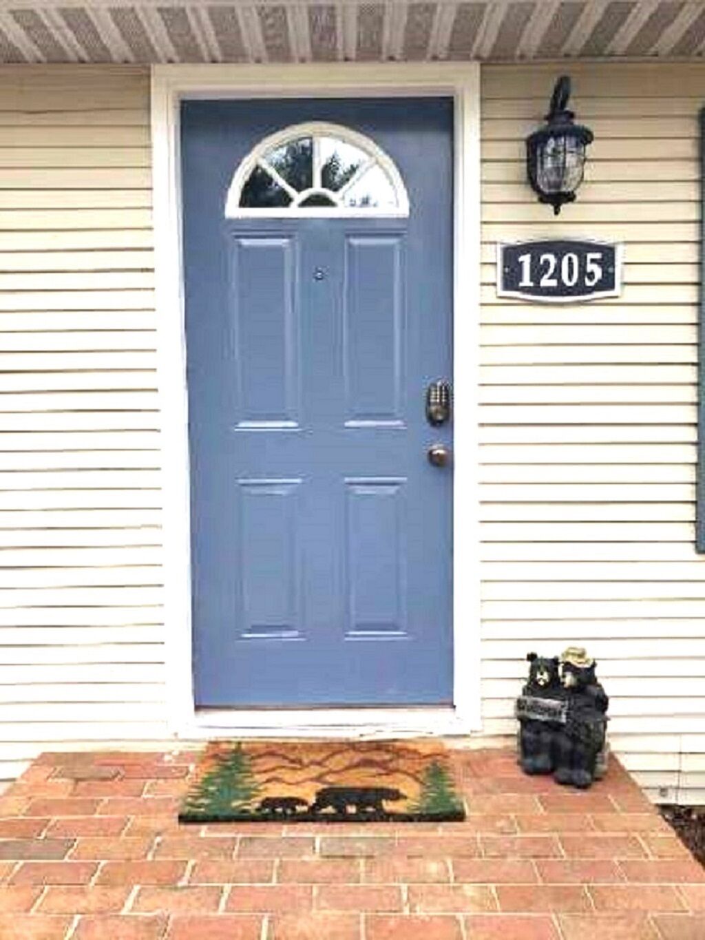 A blue door with a white number plate reading "1205" above it, a small bear figurine on the front steps, and a decorative mat with a bear design.