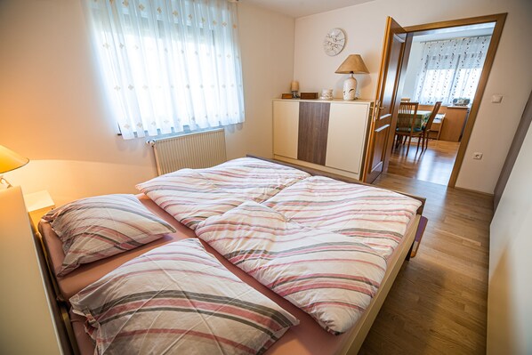 A bedroom with a bed, a radiator, a lamp, a clock, and a view into another room with a table and chairs.