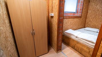A small, enclosed room with a wooden wardrobe, a window with a brick frame, and a bed with a mattress and pillows.