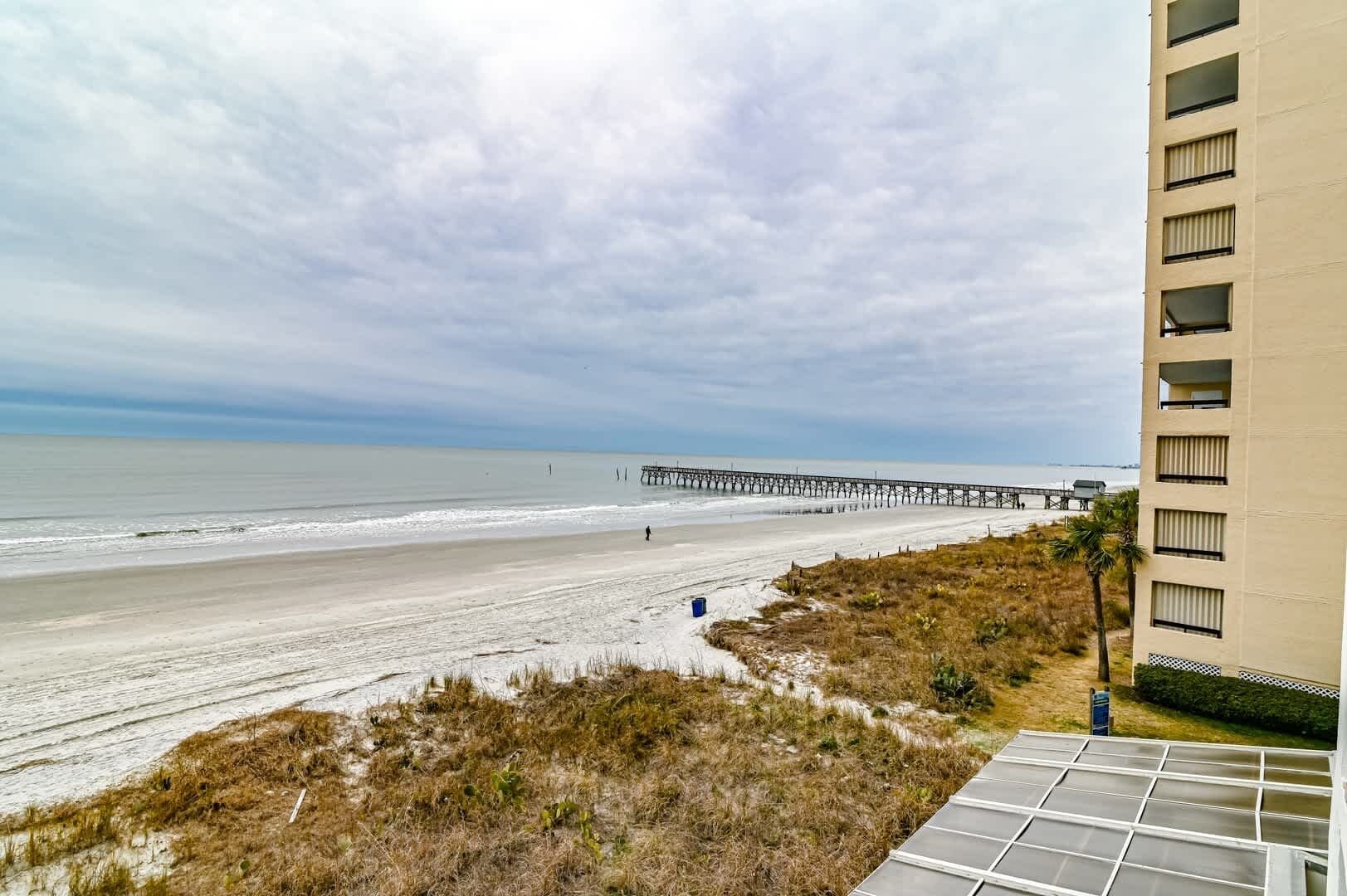 Ein Strand mit einem Pier, der sich ins Meer erstreckt, ein sandiger Uferstreifen und ein Gebäude mit vielen Fenstern.
