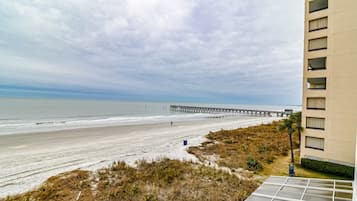 Ein Strand mit einem Pier, der sich ins Meer erstreckt, ein sandiger Uferstreifen und ein Gebäude mit vielen Fenstern.