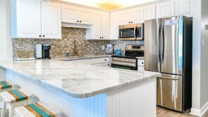 A modern kitchen with white cabinets, a stainless steel refrigerator, a microwave, and a coffee maker. - Direct Oceanfront Magic at Sea Marsh w Pool (North Myrtle Beach)