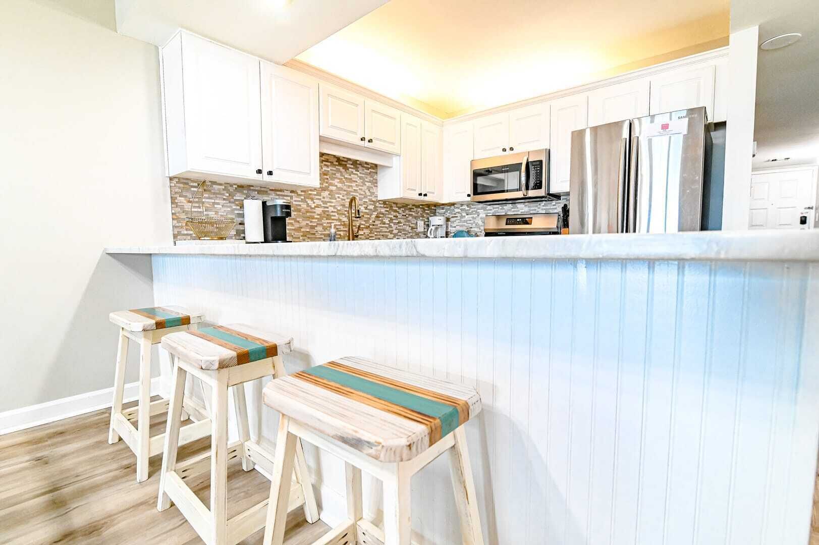 Eine Küche mit weißen Schränken, einer Stein-Spritzschutzwand, einer Mikrowelle, einem Kühlschrank und einer Bar mit Hockern.