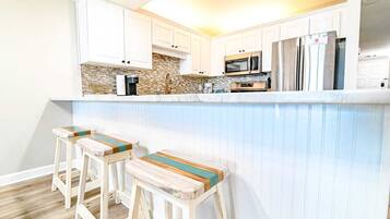 Eine Küche mit weißen Schränken, einer Stein-Spritzschutzwand, einer Mikrowelle, einem Kühlschrank und einer Bar mit Hockern.