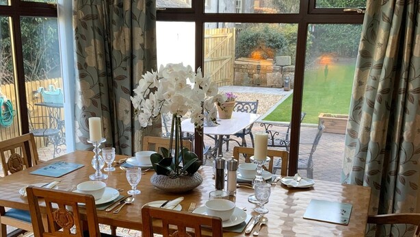 A dining area with a table set for four, surrounded by large windows with curtains. - Remarkable 2-bed Cottage in Tranquil Setting (Wooler)