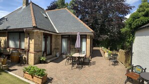 A stone house with a tiled roof, a patio with a table and chairs, and a garden with potted plants. - Remarkable 2-bed Cottage in Tranquil Setting (Wooler)