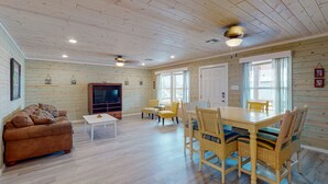 Un salon spacieux avec un plafond en bois, un canapé, un téléviseur, une table à manger avec des chaises et une fenêtre avec des rideaux.