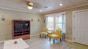 Une pièce aux murs en bois, avec une grande télévision, deux fauteuils à motifs, une petite table et une porte.