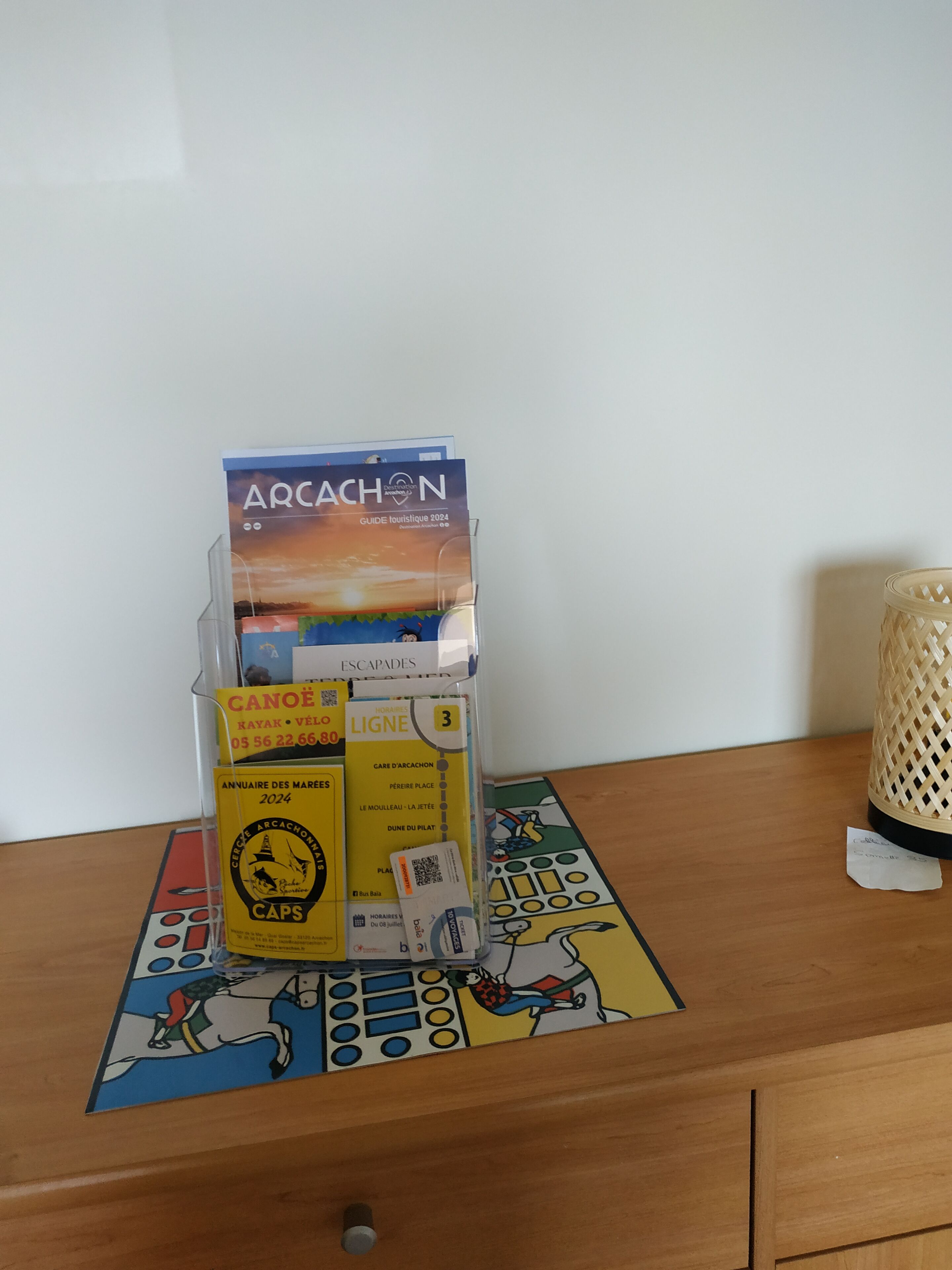 A wooden table with a board game, a brochure titled "ARCACHON GUIDE TOURISTIQUE 2024", and a small basket.