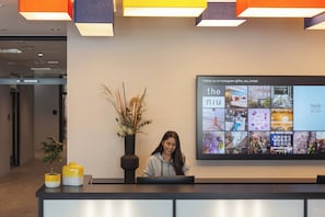 A reception desk with a digital display above it, a potted plant, and a yellow vase. - Holiday Inn - the niu, Form Stuttgart Feuerbach by IHG (Stuttgart)