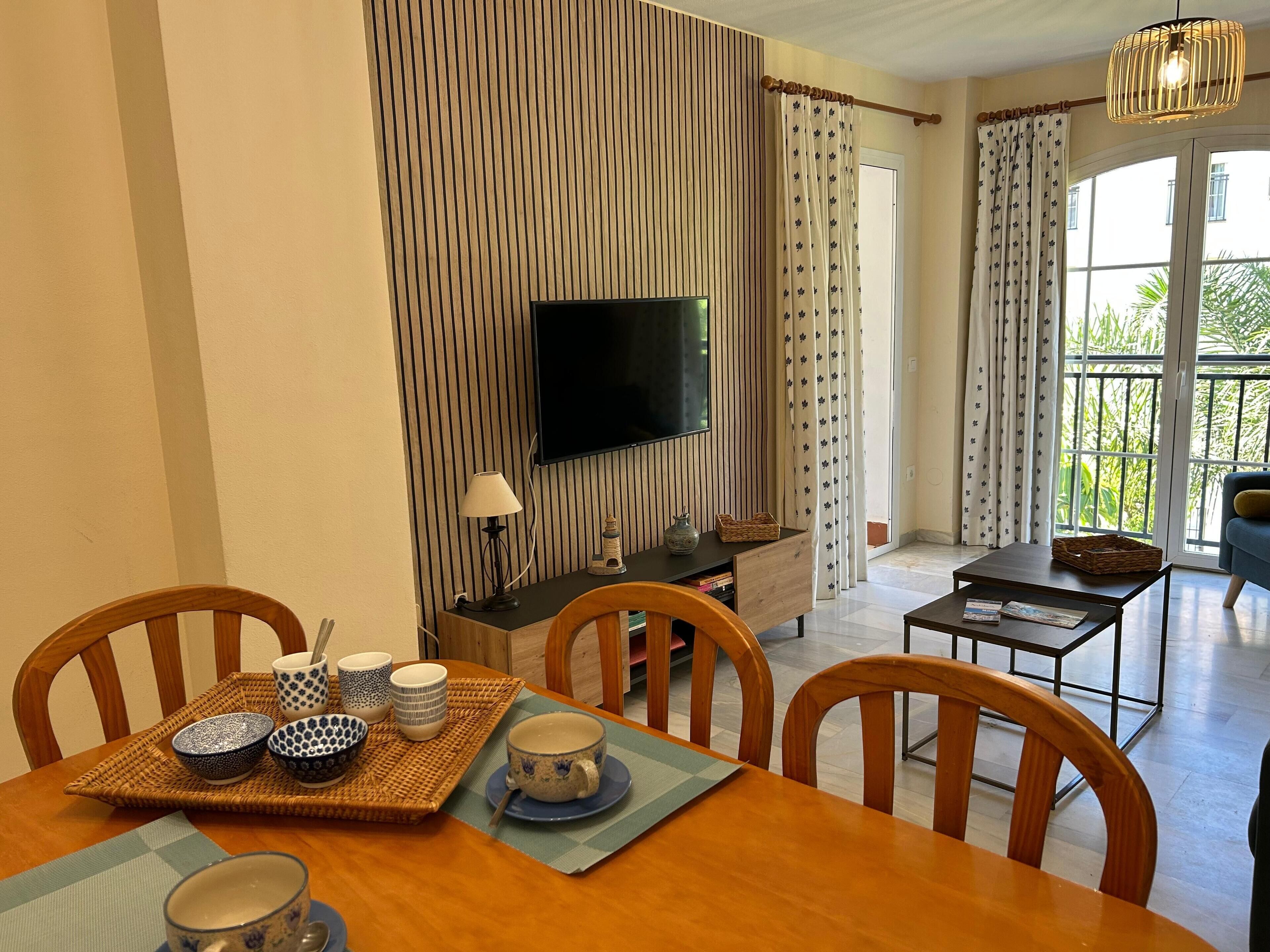 A dining area with a wooden table set for two, a television on the wall, and a balcony with curtains.