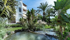 Un jardin luxuriant avec un petit étang, des palmiers et un bâtiment blanc doté de balcons.