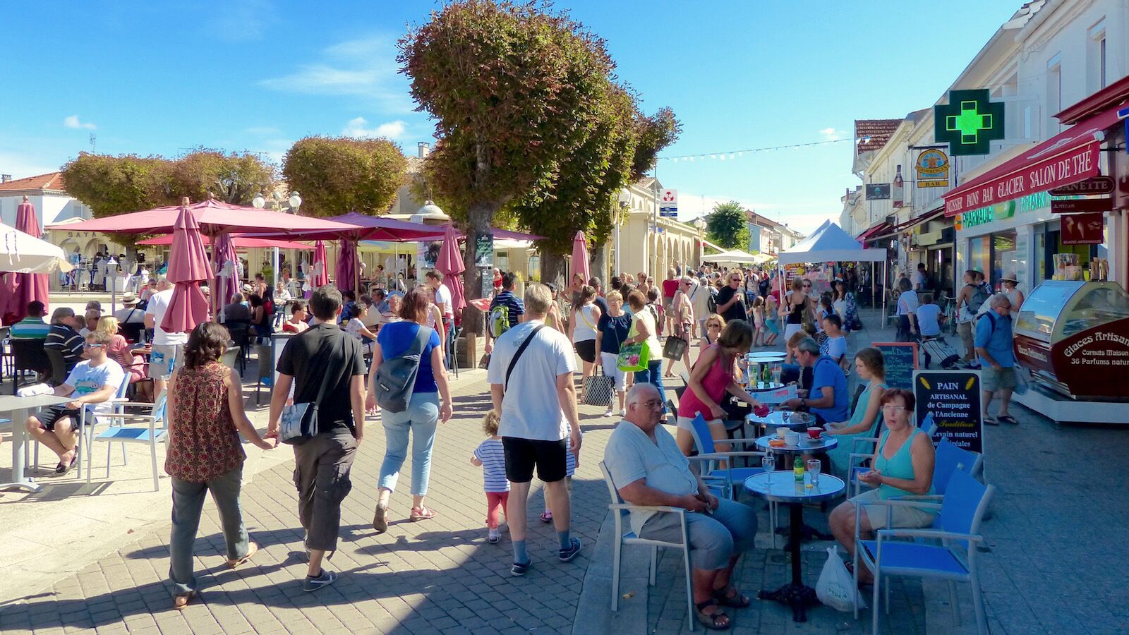 A bustling outdoor café with tables and chairs, surrounded by pedestrians and various shops.