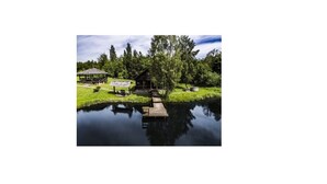 En rustik hytte ved en rolig sø med en træbro, omgivet af frodig vegetation og træer.