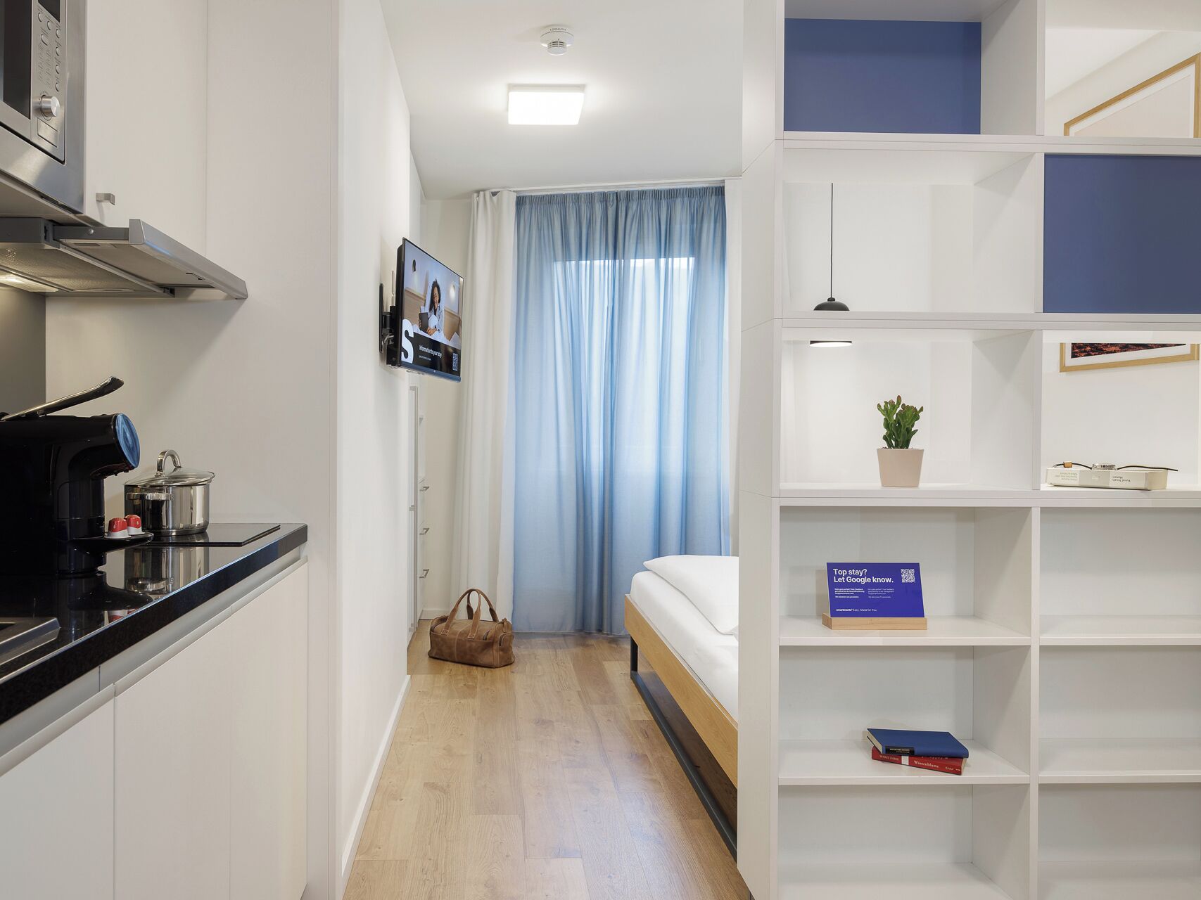 A compact hotel room with a kitchenette, a bed, a small seating area, and a bookshelf.