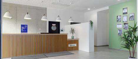 A reception area with a wooden reception desk, pendant lighting, a green wall with framed pictures, and a potted plant.