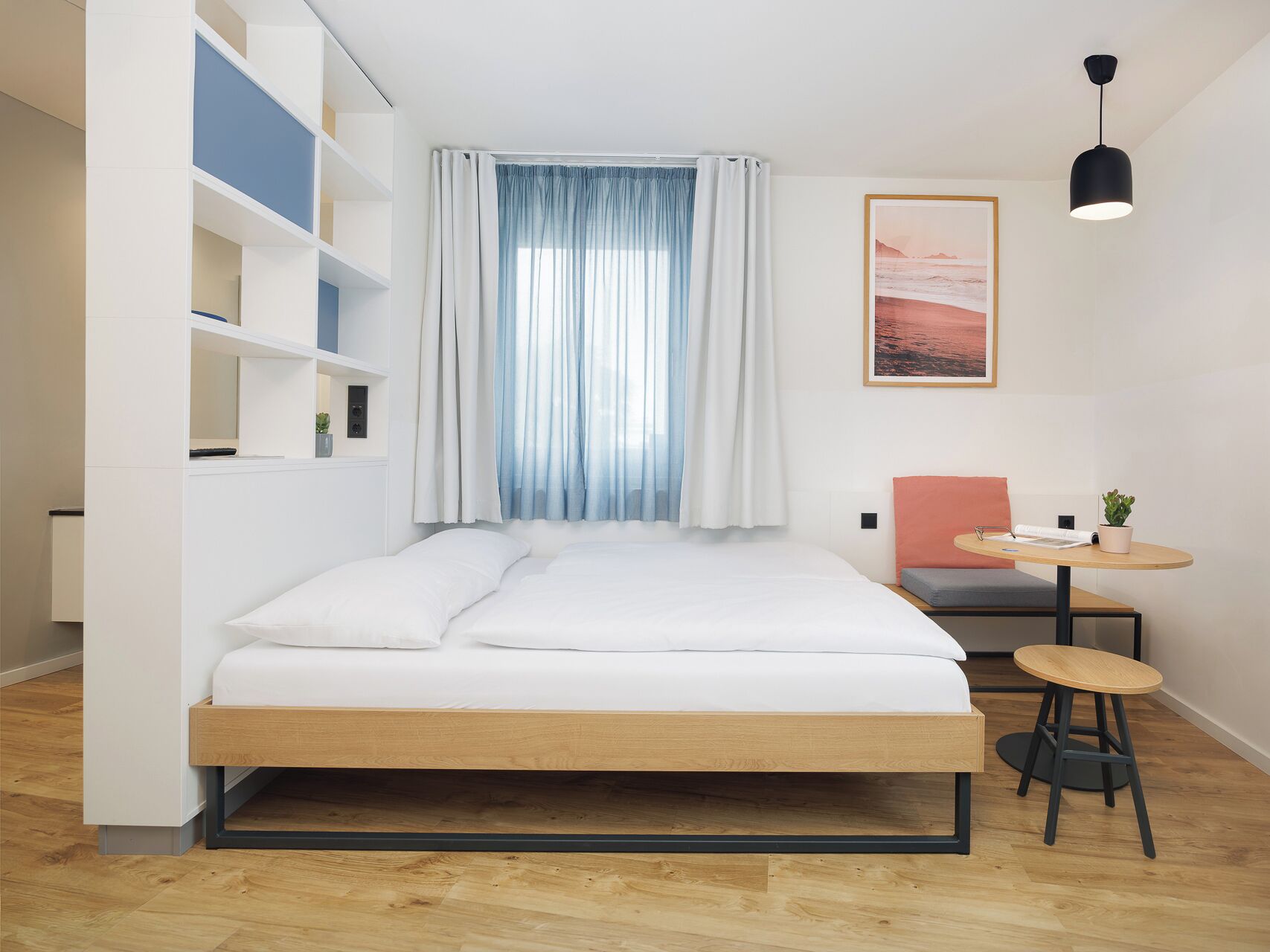 A modern bedroom with a bed, a small table, a chair, a potted plant, and a painting on the wall.