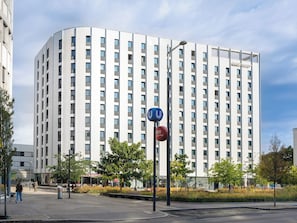 A modern high-rise building with a curved facade, multiple windows, and a pedestrian area with trees and benches. - smartments Wien Heiligenstadt (Vienna)