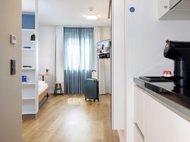 A hotel room with a bed, a small table with a plant, a suitcase, a television, and a kitchenette area.