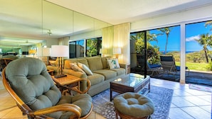 A modern living room with a sofa, chairs, and a glass coffee table. There is a view of the ocean through large windows.