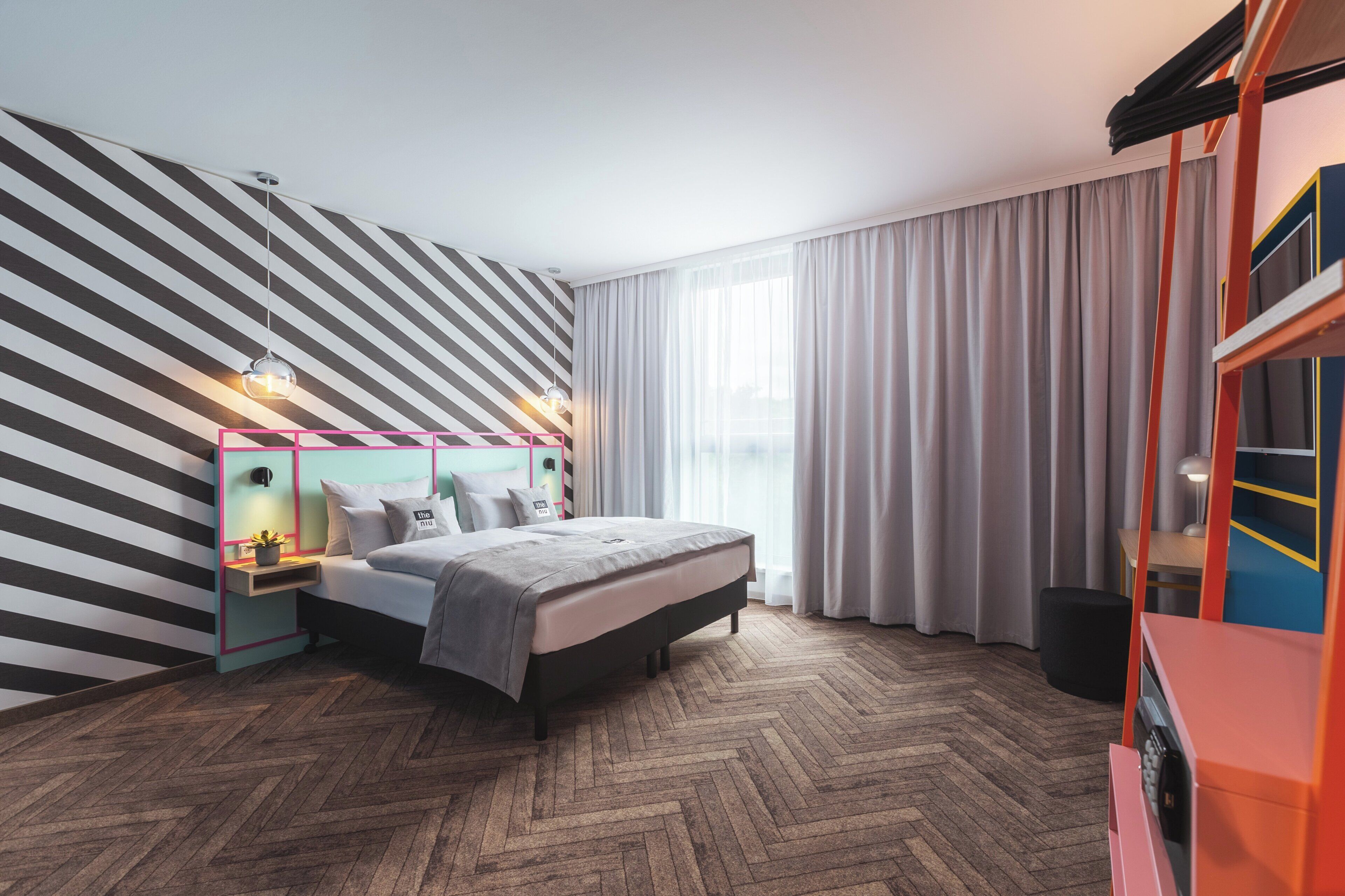 A modern bedroom with a herringbone wood floor, a bed with a grey headboard, a nightstand with a lamp, and a large window with sheer curtains.