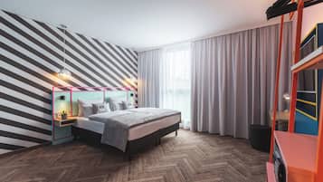 A modern bedroom with a herringbone wood floor, a bed with a grey headboard, a nightstand with a lamp, and a large window with sheer curtains.