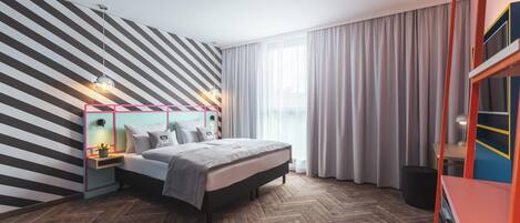 A modern bedroom with a herringbone wood floor, a bed with a grey headboard, a nightstand with a lamp, and a large window with sheer curtains.