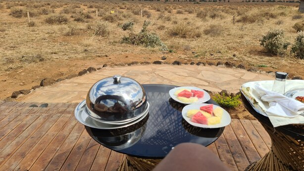 A dining table with a view of a dry landscape, a covered pot, and plates with watermelon slices. - Amanya 1 -bed King Lion Tent (Amboseli)