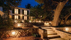 Una escalera de piedra conduce a una casa de fachada pétrea y múltiples ventanales, rodeada de árboles e iluminación exterior.